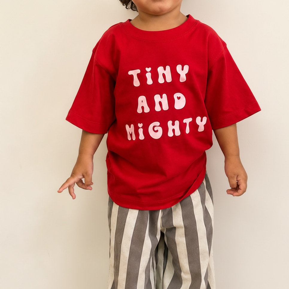 A toddler wearing a bright red short-sleeve t-shirt with 'TINY AND MIGHTY' printed in light pink bubble letters, paired with gray and white vertically striped pants, standing against a plain off-white wall.