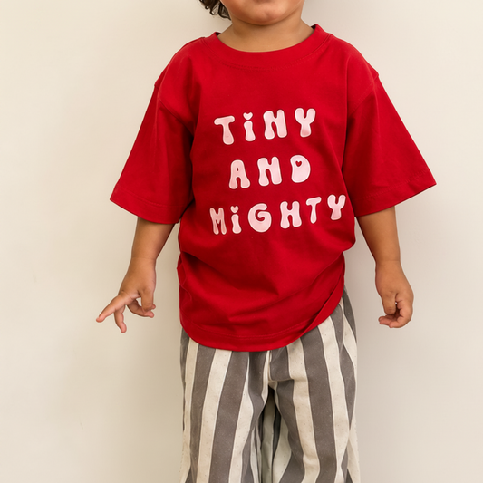 A toddler wearing a bright red short-sleeve t-shirt with 'TINY AND MIGHTY' printed in light pink bubble letters, paired with gray and white vertically striped pants, standing against a plain off-white wall.