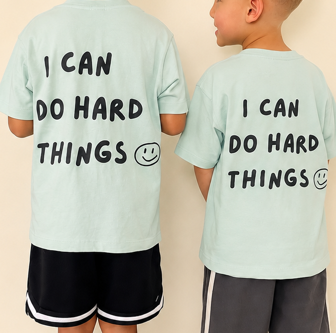 Two children standing with their backs to the camera wearing matching light mint green t-shirts with the black printed text 'I CAN DO HARD THINGS' and a smiley face on the back, against a solid beige background. Very sharp and clear image
